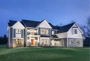 A large two-story house with a mix of dark siding and white trim, illuminated by exterior lights at twilight.