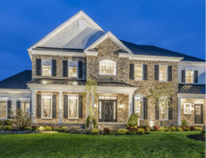Two-story stone house with white trim, numerous windows, and a manicured lawn, illuminated by exterior lights at dusk.
