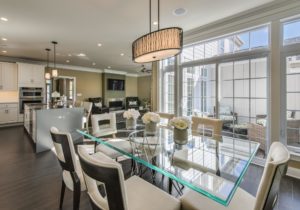 Modern kitchen and dining area with a glass table, beige chairs, and a view of the adjoining living room, featuring large windows and contemporary decor.