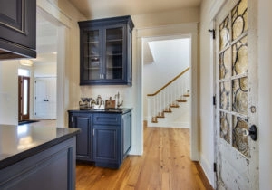 Modern kitchen with blue cabinets and a wooden floor, next to a hallway with a staircase and a decorative door with glass panels.