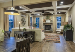 A cozy living room with wooden floors, a stone fireplace, and white sofas. Beige curtains frame the windows, and a colorful rug lies in front of the fireplace. A dining table is in the foreground.