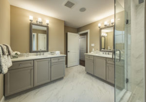 A modern bathroom features two gray vanities with mirrors, a glass-enclosed shower, beige walls, and a white marble floor.