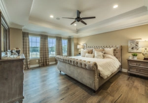A spacious bedroom with a large upholstered bed, wooden nightstands, curtains, and a ceiling fan. Decor includes lamps, flowers, and decorative items on a dresser.