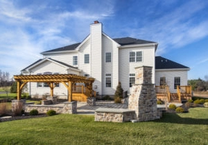 A spacious two-story white house with a wooden pergola, deck, and stone patio in a well-maintained backyard under a blue sky.