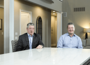 Two men sit at a marble table in a modern, warmly-lit room. The man on the left wears a suit; the man on the right wears a light blue shirt. They are smiling and appear to be in conversation.