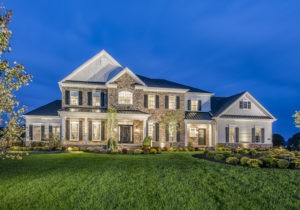 A two-story house with stone and white siding exterior, large windows, and a well-manicured lawn is illuminated at dusk.