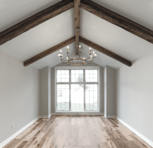 A bright, empty room with a large window, wooden ceiling beams, a chandelier, and light wood flooring.