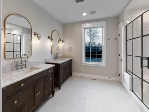 A clean and modern bathroom with two sinks, marble countertops, wooden cabinets, large mirrors, wall-mounted lights, a window, and a glass-enclosed shower.