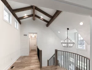 A modern hallway with wooden ceiling beams, white shiplap walls, and wood floors. There is a staircase with a wrought iron railing and a chandelier hanging from the ceiling.