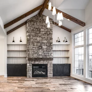 Living room with a vaulted ceiling, exposed wooden beams, stone fireplace, built-in shelves, and large windows. Chandelier hanging from the ceiling.