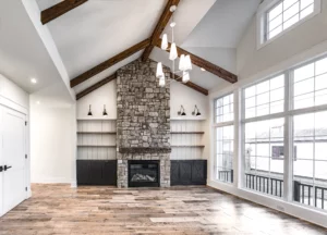 A living room with a vaulted ceiling, exposed wooden beams, and large windows. It features a stone fireplace, built-in shelves, and hardwood floors. A modern chandelier hangs from the ceiling.