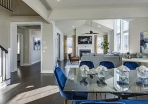 A modern open-plan living room and dining area with a glass dining table, blue chairs, a white sectional sofa, and a fireplace beneath a mounted TV.