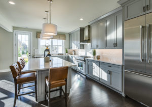A modern kitchen with gray cabinets, stainless steel appliances, a large island with a white countertop, two brown leather stools, and pendant lighting. A window and door are visible in the background.