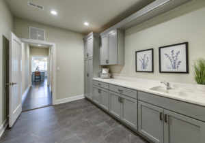 A modern laundry room with grey cabinets, a sink, a washing machine, and framed botanical art on the wall. The floor is tiled, and there's a hallway leading to another room.