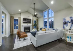 Modern living room with a light gray sectional sofa, two brown chairs, a fireplace, and a wall-mounted TV. Large windows allow natural light, and a ceiling fan hangs in the center of the room.