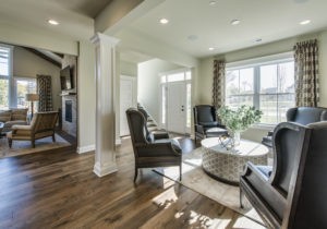 Modern living room with hardwood floors, black armchairs around a circular coffee table, a fireplace, and large windows allowing natural light. Stairs and a white front door are visible in the background.