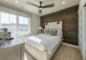 A modern bedroom with a wooden accent wall, white bed, and a large window letting in natural light. There is a ceiling fan, a bedside lamp, and a dresser in the corner.