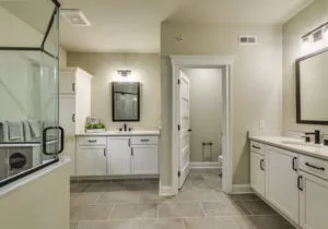 Modern bathroom with light beige walls, tiled floor, glass-enclosed shower, double sinks with large mirrors, and white cabinetry. A separate room contains a toilet.